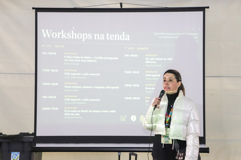 Apresentadora com microfone diante da projeção da programação de workshops.