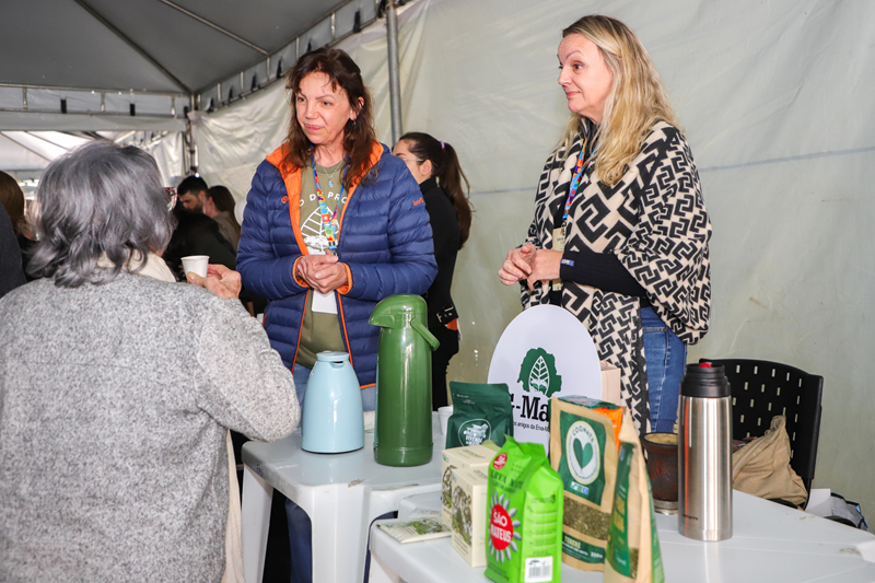 Visitantes conversam com expositoras junto a garrafas térmicas e pacotes de erva-mate.